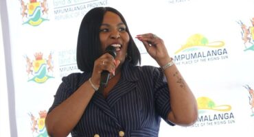 a woman speaks over a a microphone. There is a backdrop with the government logos