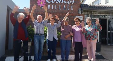 old people with hands in the air in front of a building named Kosmos Village