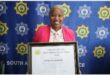 A woman in a pink jacket holds up an accolade. behind her is a police SAPS backdrop