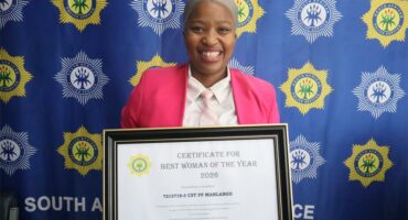 A woman in a pink jacket holds up an accolade. behind her is a police SAPS backdrop