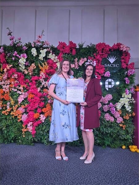 two women holding up an award. behind them is a large flower wall display