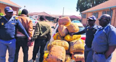 police with a huge load of bags. two men are hand cuffed in the background