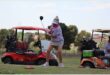 Woman with a white cap whacks the ball. There are golf carts in the background